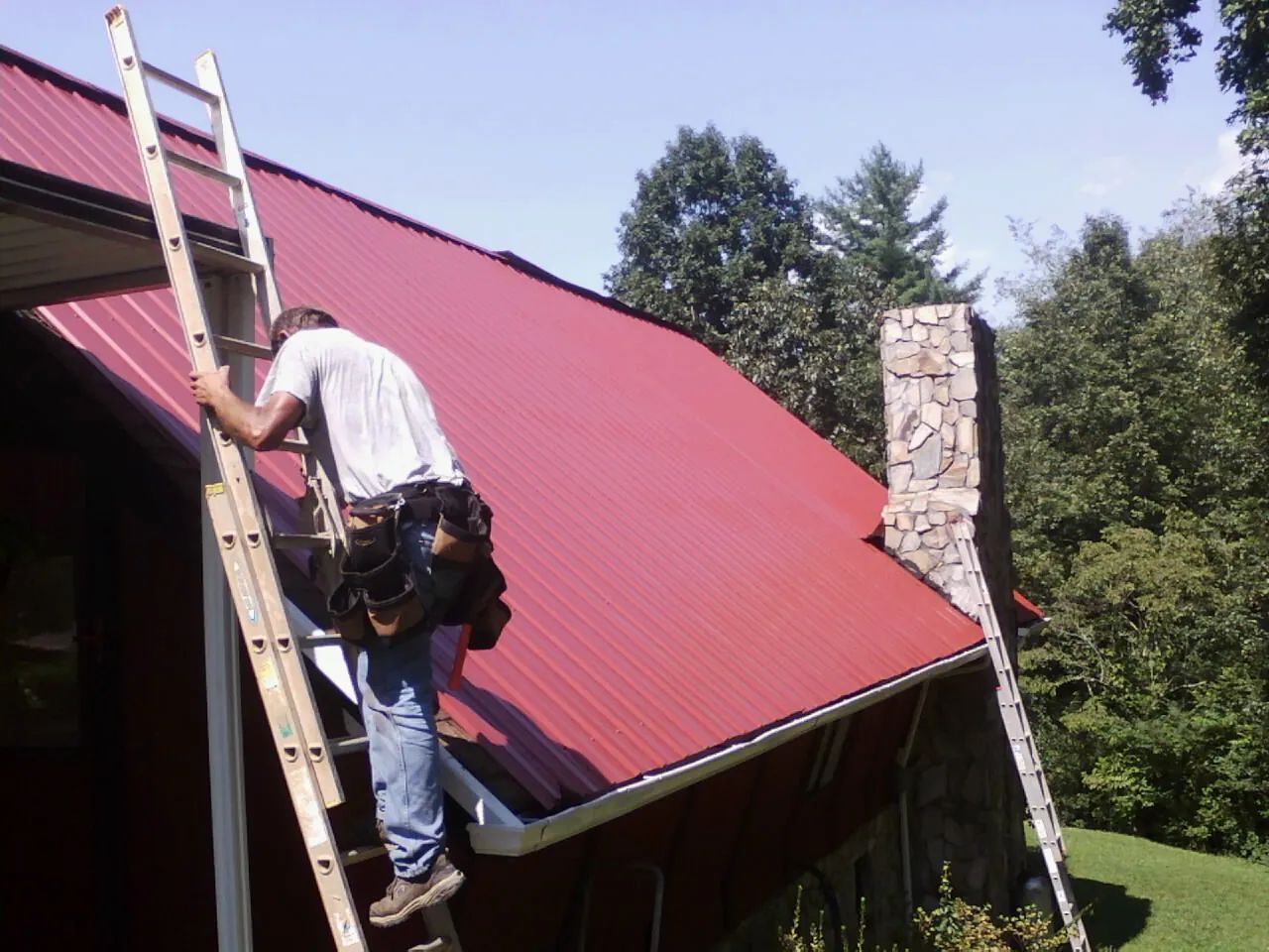 Roofer on a ladder beside a red metal roof and stone chimney — Soffit & Fascia Repair by Roofing Brothers in Maple Heights